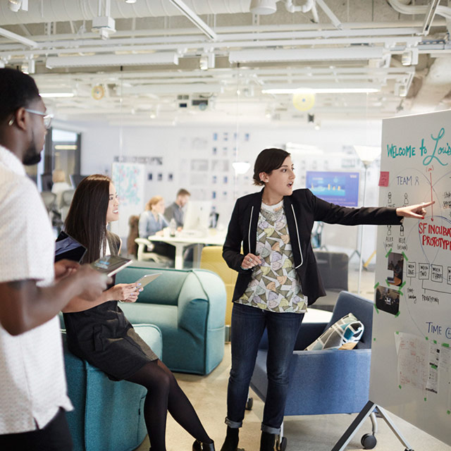 Woman pointing to a whiteboard while two people watch.