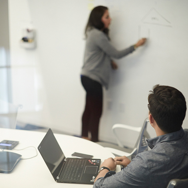 Woman writing on a whiteboard and a man looking at her while sitting at a desk with his laptop open.