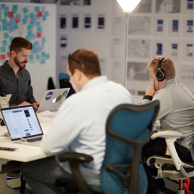 Three men sitting around a desk while looking at their laptop.